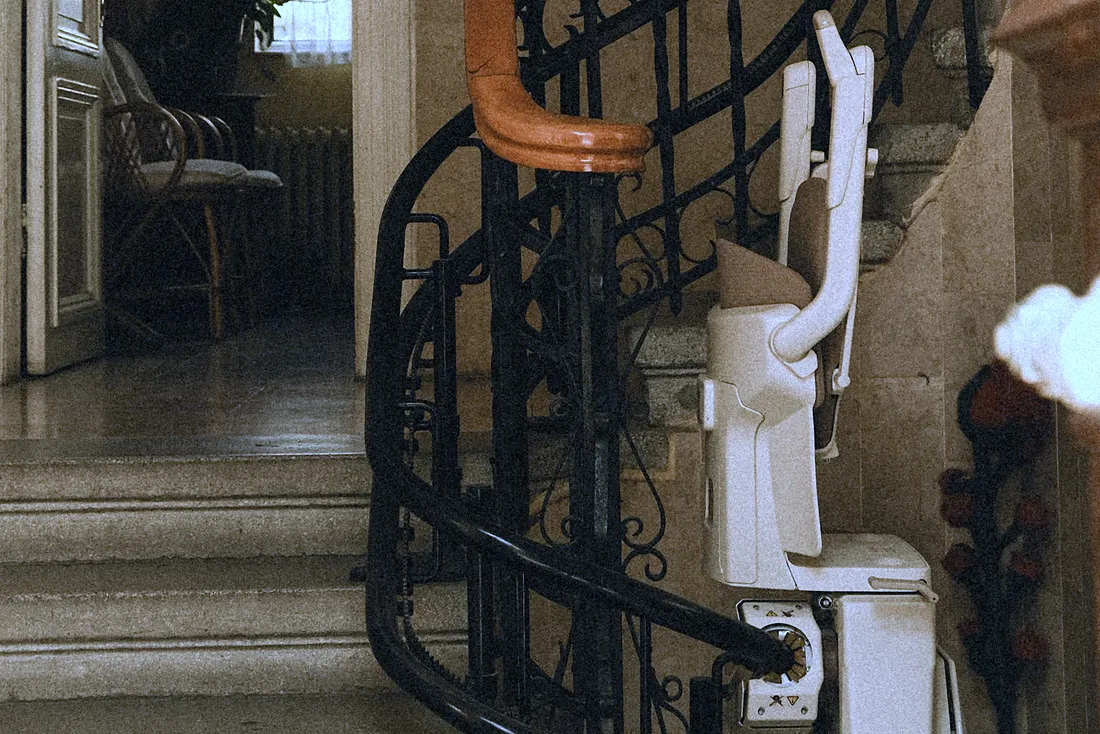 Siège de monte-escalier replié au pied d'un escalier tournant à rampe en fer forgé, dans le hall d'une maison ancienne (Aspach, Haut-Rhin)