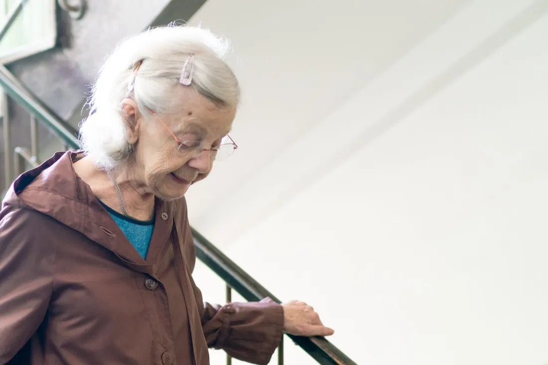 Femme âgée aux cheveux blancs qui descend une marche, la main sur la rampe (Caudecoste, Lot-et-Garonne)