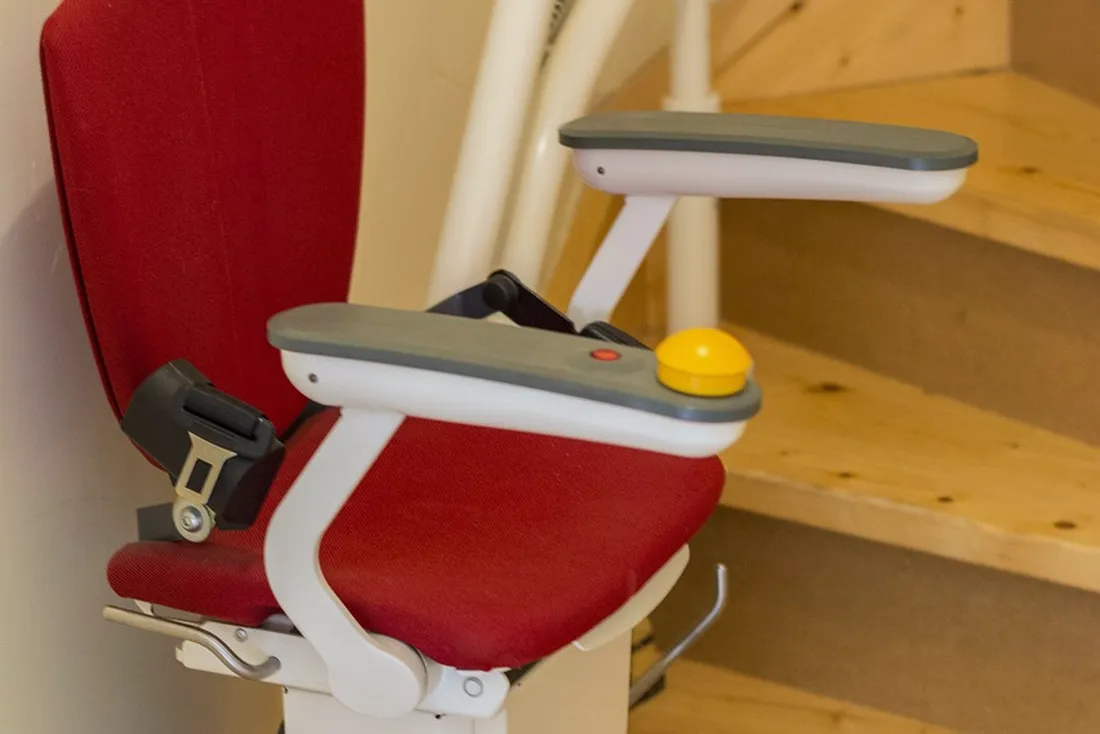 Fauteuil de monte-escalier à assise rouge, ceinture de sécurité et bouton de commande jaune sur l'accoudoir, au bas d'un escalier en bois clair