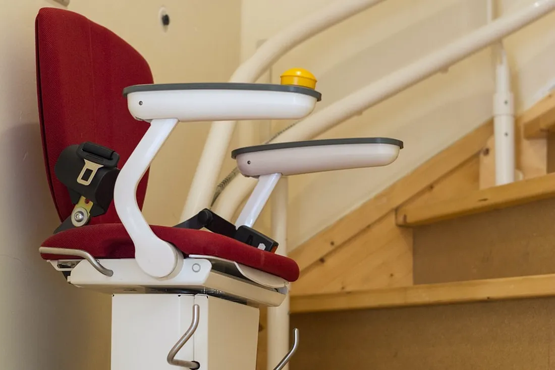 Fauteuil rouge de monte-escalier vu de côté, accoudoirs blancs, sur un rail blanc cintré qui suit un escalier tournant en bois (Doue, Seine-et-Marne)